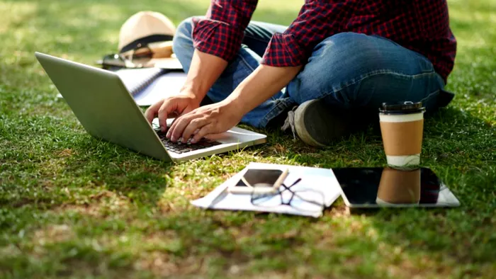 Un băiat care stă pe iarbă și tastează pe laptop.