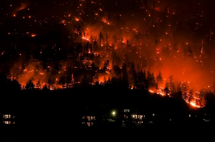 Flăcările de la incendiul forestier McDougall Creek înconjoară o panta de munte deasupra caselor din West Kelowna, British Columbia, pe 18 august. Condițiile meteorologice extreme au alimentat cea mai gravă sezon de incendii din istoria Canadei, cu mii de incendii devastând milioane de acri și întunecând cerul la mii de mile distanță. [Fotograf: Darryl Dyck—The Canadian Press/AP]